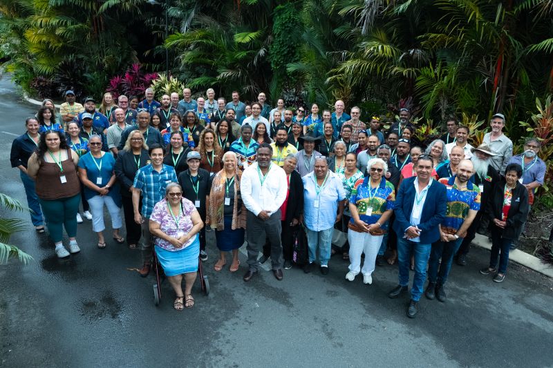 Large group of smiling Traditional Owners with tropical plants in the background.