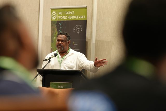 A male Indigenous leader with black and grey hair stands at a lectern delivering a presentation, hand raised.